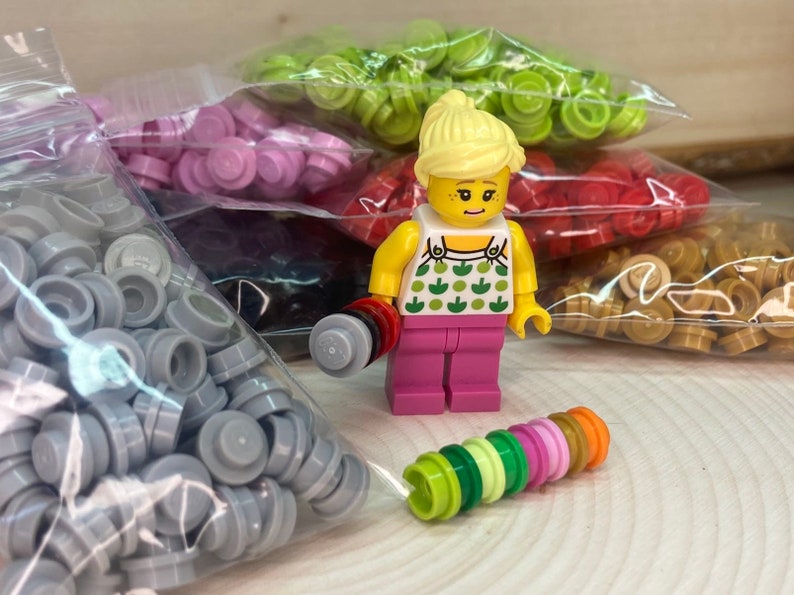May include: A small plastic figurine of a person wearing a pink shirt with green apples on it. The figurine is surrounded by bags of small plastic pieces in various colors, including gray, pink, green, red, and gold.