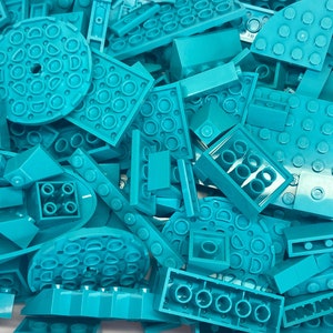 May include: A pile of turquoise building blocks in various shapes and sizes. The blocks include flat squares, rectangular bricks, and circular pieces with stud details. The pieces are scattered, creating a textured and playful arrangement.