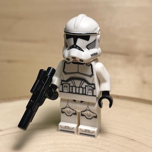May include: A white Lego Star Wars Clone Trooper minifigure with black detailing and a black blaster rifle. The figure has a white helmet and armor with black accents. The toy is standing on a wooden surface.