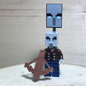 May include: A Minecraft-themed toy figure of a Pillager, featuring a gray block head with pixelated eyes and a brown crossbow. The figure wears a brown jacket, blue pants, and stands on a wooden surface. The figure has a second head on top.