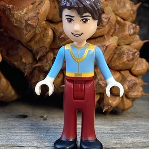 May include: A small plastic figurine of a boy wearing a blue shirt with a gold chain, red pants, and black shoes.