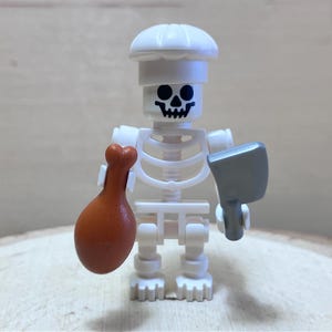 May include: A white skeleton figure wearing a chef's hat, holding a brown drumstick in one hand and a gray cleaver in the other. The figure has a black skull face and is made of plastic. The figure is standing on a wooden surface.