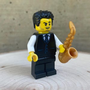 May include: A Lego musician minifigure holding a gold saxophone. The figure has black hair, a yellow face with a goatee, and wears a black waistcoat over a white shirt and black trousers. The saxophone is a curved design.