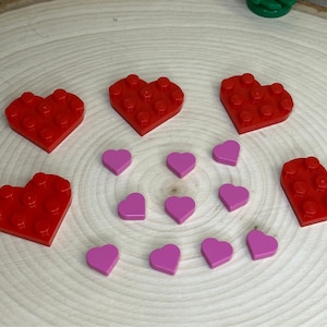 Può includere: Una collezione di mattoncini LEGO a forma di cuore rosso e rosa. I cuori rossi sono più grandi e hanno una superficie testurizzata. I cuori rosa sono più piccoli e lisci.