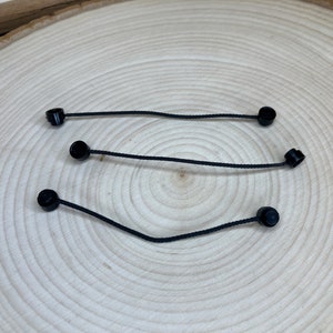 May include: Three black cord stoppers with a black cord attached to each. The stoppers are round and have a smooth surface. The cords are thin and flexible.