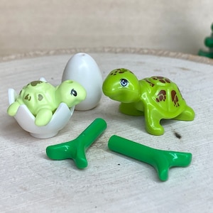 Może przedstawiać: Zestaw figurek żółwi. Jeden żółw wychodzi z pękniętej białej skorupki jajka, drugi stoi, a trzeci to zielona gałązka. Żółwie są jasnozielone z brązowymi plamkami.