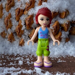 Puede incluir: Una figura de Lego de una ni&ntilde;a con cabello rojo, que lleva un top azul sin mangas con dise&ntilde;os de mariposas y huellas de patas, pantalones cortos verdes con dise&ntilde;os de mariposas y sandalias moradas. La figura est&aacute; de pie sobre una superficie marr&oacute;n con nieve.
