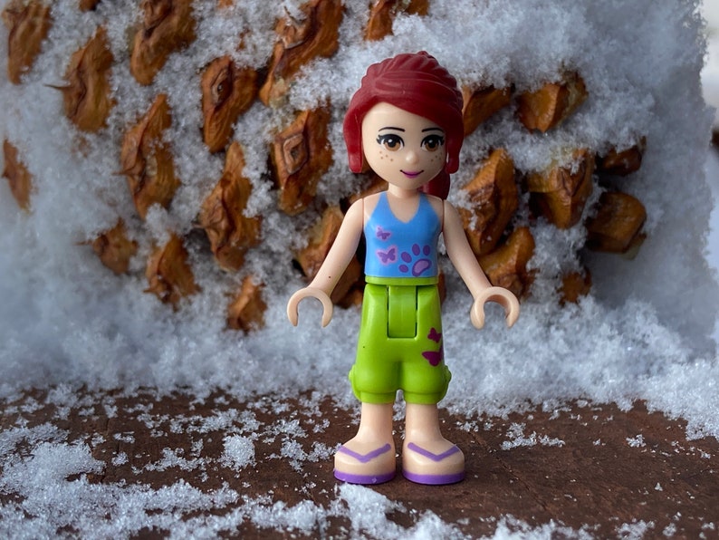 Puede incluir: Una figura de Lego de una ni&ntilde;a con cabello rojo, que lleva un top azul sin mangas con dise&ntilde;os de mariposas y huellas de patas, pantalones cortos verdes con dise&ntilde;os de mariposas y sandalias moradas. La figura est&aacute; de pie sobre una superficie marr&oacute;n con nieve.