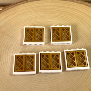 May include: Five white Lego window pieces with gold-colored lattice designs. The windows are arranged on a light brown, circular wooden surface. The windows are a building block toy.
