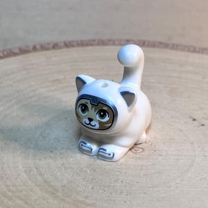 May include: White cat figurine with brown eyes and a silver nose. The cat is sitting with its tail curled up behind it.