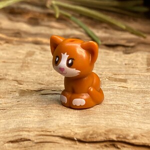 May include: A small orange and white cat figurine with a white nose and pink cheeks. The cat is sitting with its paws tucked under its body.