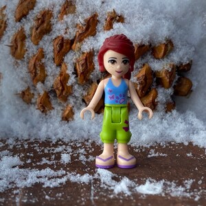Puede incluir: Una peque&ntilde;a mu&ntilde;eca de pl&aacute;stico con cabello rojo, un top azul y morado, pantalones verdes con un dise&ntilde;o de huella de pata y sandalias moradas. La mu&ntilde;eca est&aacute; de pie sobre una superficie marr&oacute;n cubierta de nieve.