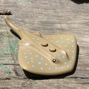 May include: A brown and tan plastic toy stingray with blue spots. The toy has two small black eyes and two round holes on the top.