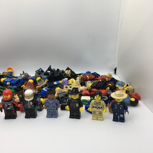 May include: A collection of assorted Lego minifigures, including police officers, construction workers, and other characters. The minifigures are arranged in a pile on a white surface.