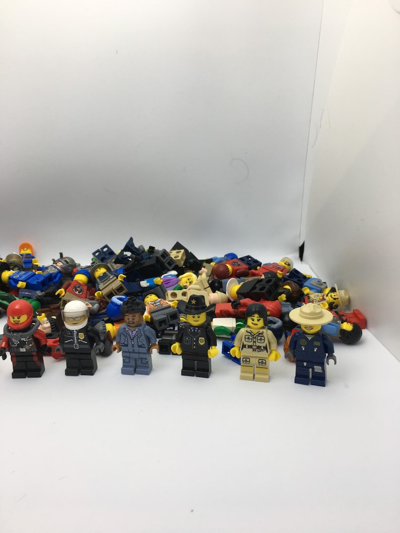 May include: A collection of assorted Lego minifigures, including police officers, construction workers, and other characters. The minifigures are arranged in a pile on a white surface.