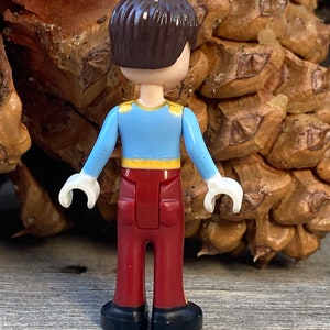 May include: A small plastic figurine of a boy wearing a blue shirt with a gold trim, red pants, and black shoes. The figurine is standing on a wooden surface.