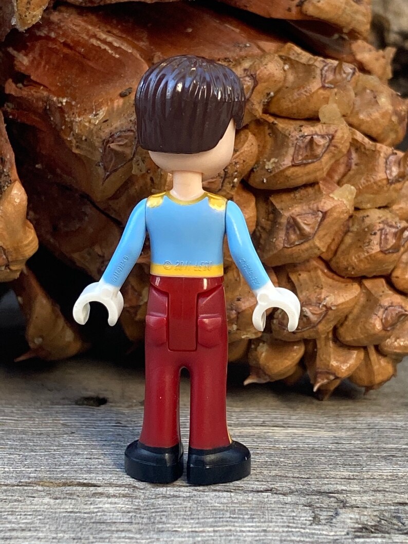 May include: A small plastic figurine of a boy wearing a blue shirt with a gold trim, red pants, and black shoes. The figurine is standing on a wooden surface.