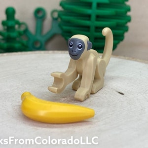 May include: A tan toy monkey with a gray face and ears reaches toward a yellow banana. The monkey is positioned in front of the banana. The background includes green plastic shapes and the text "BricksFromColoradoLLC".