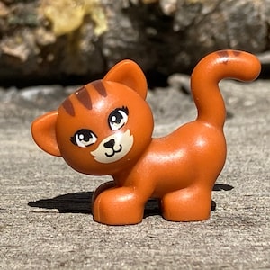 May include: A small orange and brown plastic toy cat with black stripes and white facial features. The cat has large eyes and a small, curled tail.