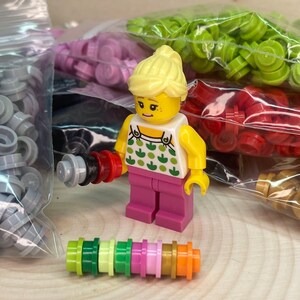 May include: A small, yellow-haired Lego figure wearing a pink dress and holding a small, multi-colored Lego tool. The figure is surrounded by bags of colorful Lego pieces.