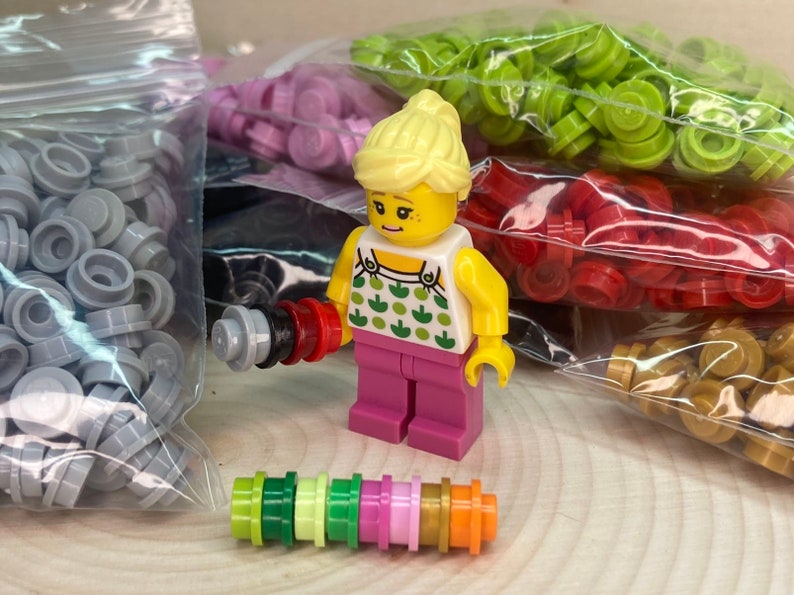 May include: A small, yellow-haired Lego figure wearing a pink dress and holding a small, multi-colored Lego tool. The figure is surrounded by bags of colorful Lego pieces.