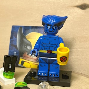 Op de afbeelding: Een blauwe Lego-figuur van Beast uit X-Men, met een gele beker met een rood-wit embleem. De figuur staat op een zwarte basis, met een Wolverine-kaart op de achtergrond. Andere Lego-accessoires zijn ook aanwezig.