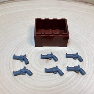 May include: A brown toy chest with six gray toy revolvers arranged around it. The chest is rectangular with a hollow interior. The revolvers are small and detailed, with visible handles and barrels. The items are on a light brown wooden surface.
