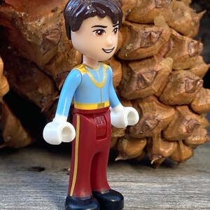 May include: A small plastic figurine of a boy wearing a blue shirt with gold trim, red pants with gold stripes, and white gloves. The figurine has brown hair and a friendly expression.