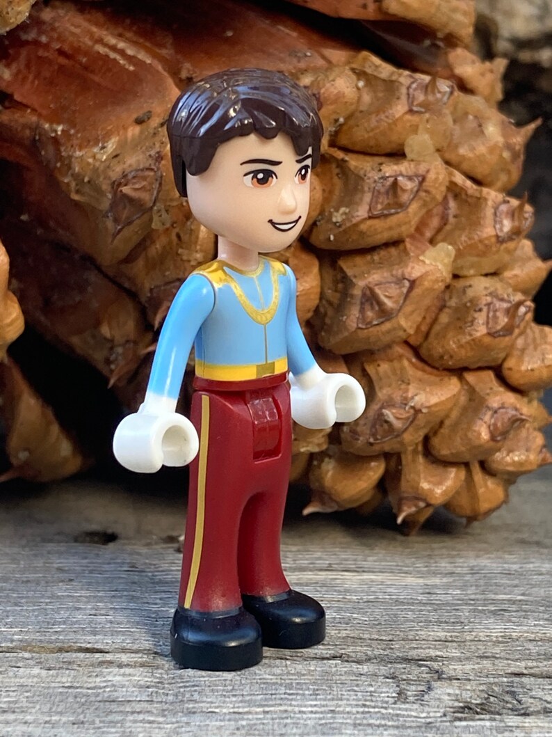 May include: A small plastic figurine of a boy wearing a blue shirt with gold trim, red pants with gold stripes, and white gloves. The figurine has brown hair and a friendly expression.