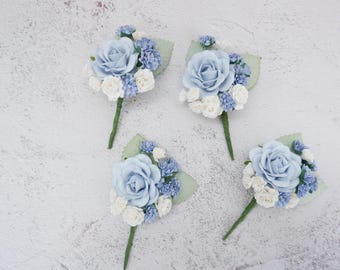 Puede incluir: Cuatro boutonnières de flores azules y blancas con una sola rosa azul y flores de relleno azules y blancas. Las boutonnières están sobre un fondo blanco.