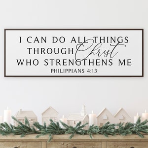 Puede incluir: Una consola de madera con un letrero enmarcado que dice "I CAN DO ALL THINGS THROUGH Christ WHO STRENGTHENS ME". La mesa está decorada con velas, casas en miniatura y una guirnalda verde. Debajo hay una cesta, una caja de madera y un arbolito.