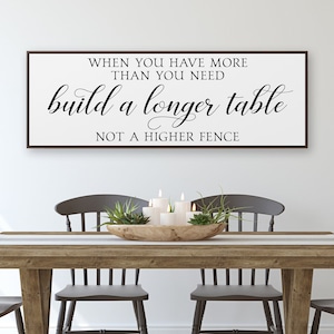 May include: A white sign with black text that reads "When you have more than you need build a longer table not a higher fence". The sign is hanging on a white wall above a wooden table with three chairs.