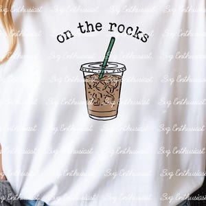 Peut inclure: T-shirt blanc avec un dessin d'un café glacé dans un gobelet en plastique transparent et une paille verte. Les mots "on the rocks" sont au-dessus de la tasse en texte noir. Le design est une illustration simple de style dessin animé.