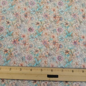 May include: A beige fabric with a floral pattern in shades of pink, blue, and brown. The fabric has a vintage, watercolor look. A ruler is placed on the fabric to show the scale.