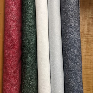 May include: A selection of fabric rolls in various colors, including burgundy, dark green, white, light gray, and navy blue. Each roll features a speckled pattern of small, lighter-colored dots. The fabrics are neatly rolled and displayed, showcasing a variety of textures and hues.