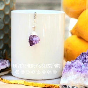 May include: A silver chain with a purple amethyst crystal pendant. The pendant is in the shape of a rough, natural crystal. The chain is hanging from a white mug with the text "LOVE ENERGY & BLESSINGS" printed on the side.