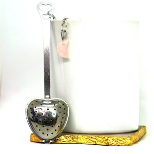 May include: Silver heart-shaped tea infuser with a rose quartz crescent moon charm attached to the handle. The infuser is sitting on a gold-colored surface.