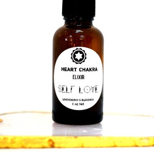 May include: A brown glass bottle with a black dropper top. The bottle has a white label with the text "Heart Chakra Elixir Self Love Love Energy & Blessings 1 oz Net".