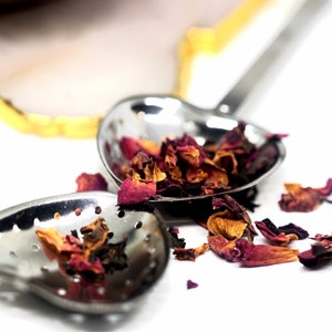 May include: Close-up of two heart-shaped stainless steel tea infusers filled with dried rose petals. The infusers are on a white surface, with some petals scattered around. The infusers have small holes for steeping tea.