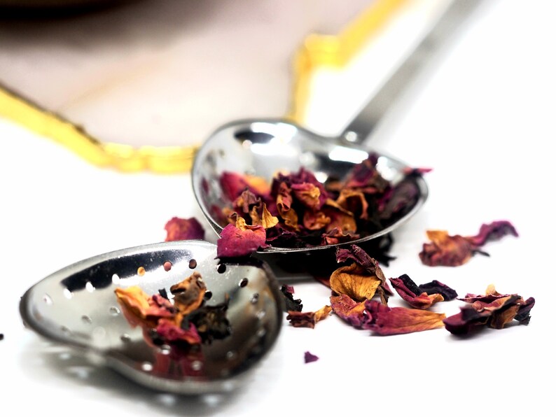 May include: Close-up of two heart-shaped stainless steel tea infusers filled with dried rose petals. The infusers are on a white surface, with some petals scattered around. The infusers have small holes for steeping tea.