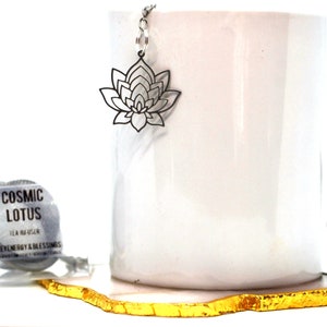 May include: A silver tea infuser shaped like a lotus flower with a chain attached. The infuser is sitting in a white mug on a gold and white coaster. The packaging for the infuser is visible in the foreground, which says "Cosmic Lotus Tea Infuser Love, Energy & Blessings".