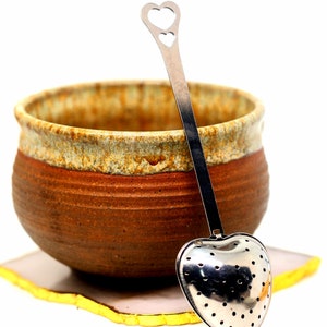 May include: A ceramic tea cup with a brown and tan exterior, paired with a heart-shaped stainless steel tea infuser. The infuser has a perforated bowl and a long handle with a heart cutout. The cup rests on a square coaster with a yellow edge.