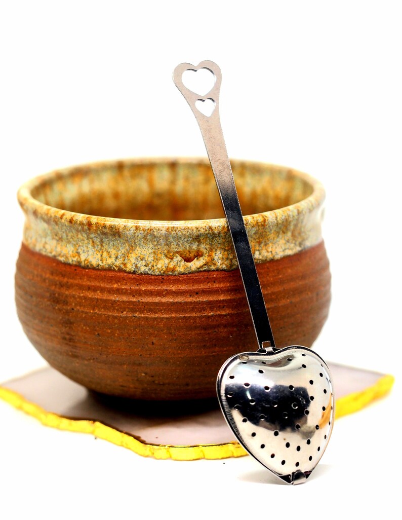 May include: A ceramic tea cup with a brown and tan exterior, paired with a heart-shaped stainless steel tea infuser. The infuser has a perforated bowl and a long handle with a heart cutout. The cup rests on a square coaster with a yellow edge.