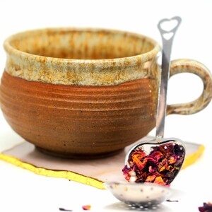 May include: A handmade ceramic mug with a brown and tan exterior, resting on a white and gold coaster. A heart-shaped tea infuser filled with dried rose petals is placed inside the mug. The mug has a rustic, artisanal appearance.