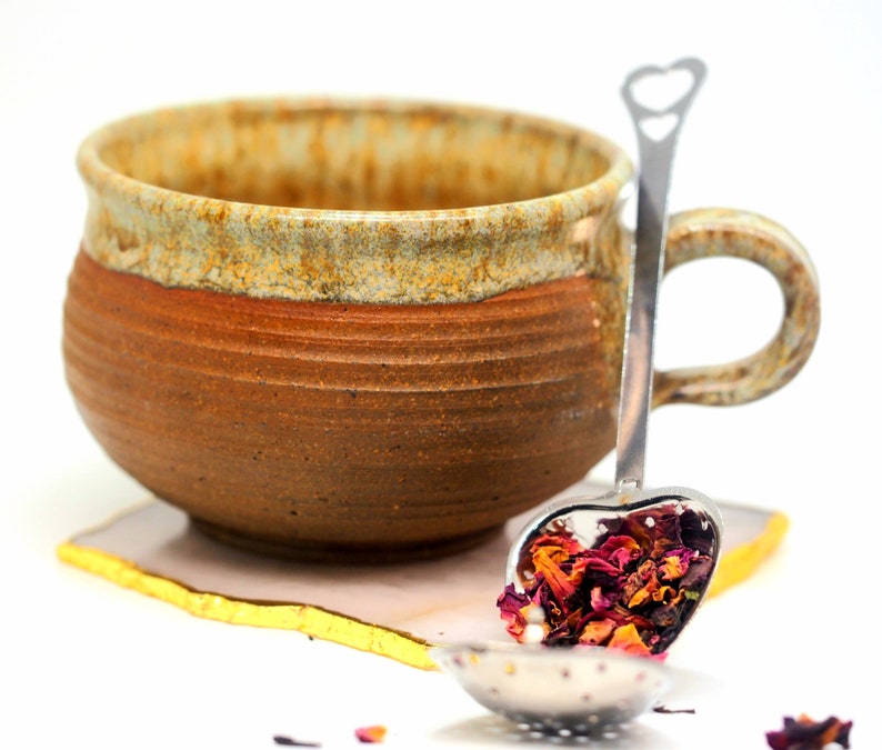 May include: A handmade ceramic mug with a brown and tan exterior, resting on a white and gold coaster. A heart-shaped tea infuser filled with dried rose petals is placed inside the mug. The mug has a rustic, artisanal appearance.