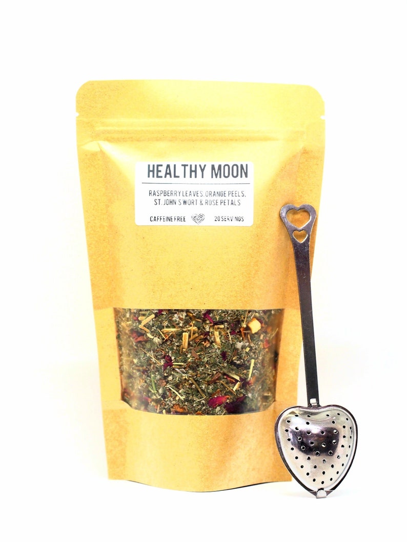 May include: A brown paper bag with a clear window showing a blend of dried herbs and flowers. The bag is labeled "HEALTHY MOON" with the text "RASPBERRY LEAVES, ORANGE PEELS, ST. JOHN SWORT & ROSE PETALS" and "CAFFEINE FREE 20 SERVINGS". A silver heart-shaped tea infuser is included.
