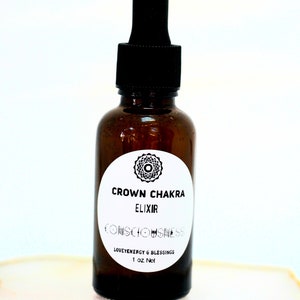 May include: A brown glass bottle with a black dropper top. The bottle has a white label that says "Crown Chakra Elixir Consciousness Love Energy & Blessings 1 oz Net".