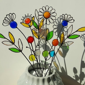 Puede incluir: Un ramo de flores de metal coloridas en un jarr&oacute;n blanco. Las flores presentan p&eacute;talos de vidrio y acentos esf&eacute;ricos en tonos azules, naranjas, amarillos, rojos y verdes. Los tallos son de metal oscuro y las hojas son de vidrieras.