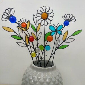 Puede incluir: Un jarr&oacute;n de cer&aacute;mica gris con un patr&oacute;n de diamantes contiene un ramo de flores decorativas de metal. Las flores presentan detalles de vidrio de colores en azul, naranja, amarillo y verde, con un dise&ntilde;o de mariposa y margarita.