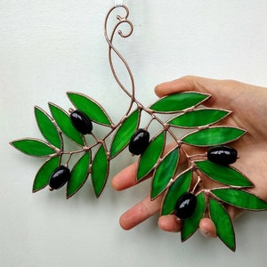 K&ouml;nnte beinhalten: Ein Olivenzweig aus Glasmalerei mit gr&uuml;nen Bl&auml;ttern und schwarzen Oliven. Die Bl&auml;tter sind ein leuchtendes Gr&uuml;n, und die Oliven sind gl&auml;nzend schwarz. Der kupferfarbene Rahmen verleiht einen Hauch von Eleganz. Ein dekoratives St&uuml;ck f&uuml;r die Inneneinrichtung.